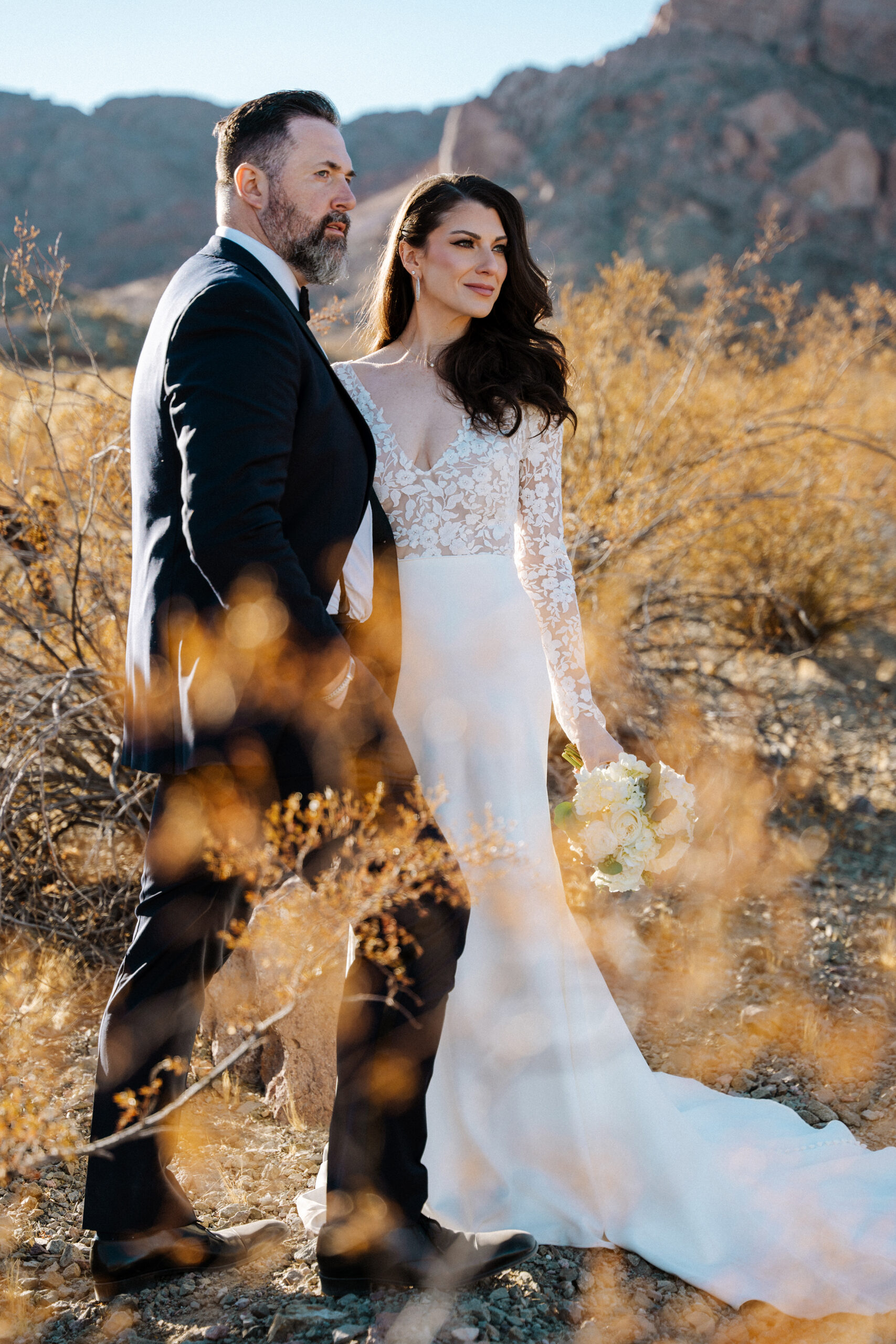 Bride and groom walking through desert brush with mountains in the background during Nelson Ghost Town wedding