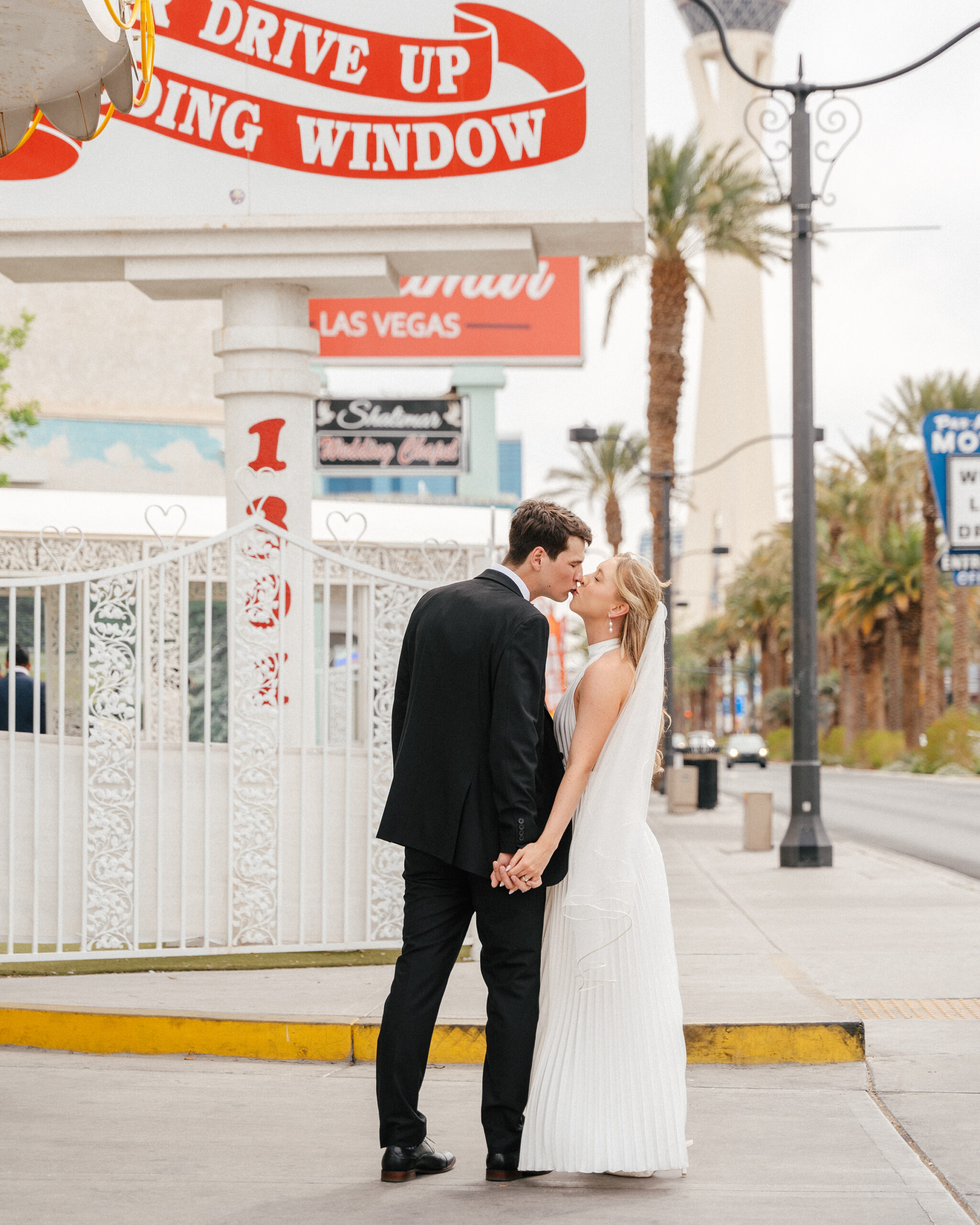 Newlyweds sharing a kiss at golden hour outside A Little White Wedding Chapel, with soft light and Vegas skyline in the background.