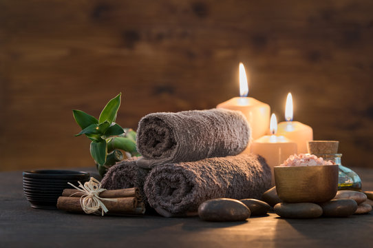 Rolled brown spa towels, candles, massage stones, and essential oils arranged on a wooden table in a relaxing spa setting.