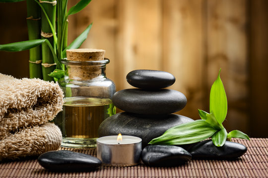 Stacked black massage stones, bamboo leaves, essential oil, a candle, and towels arranged in a calming spa environment.