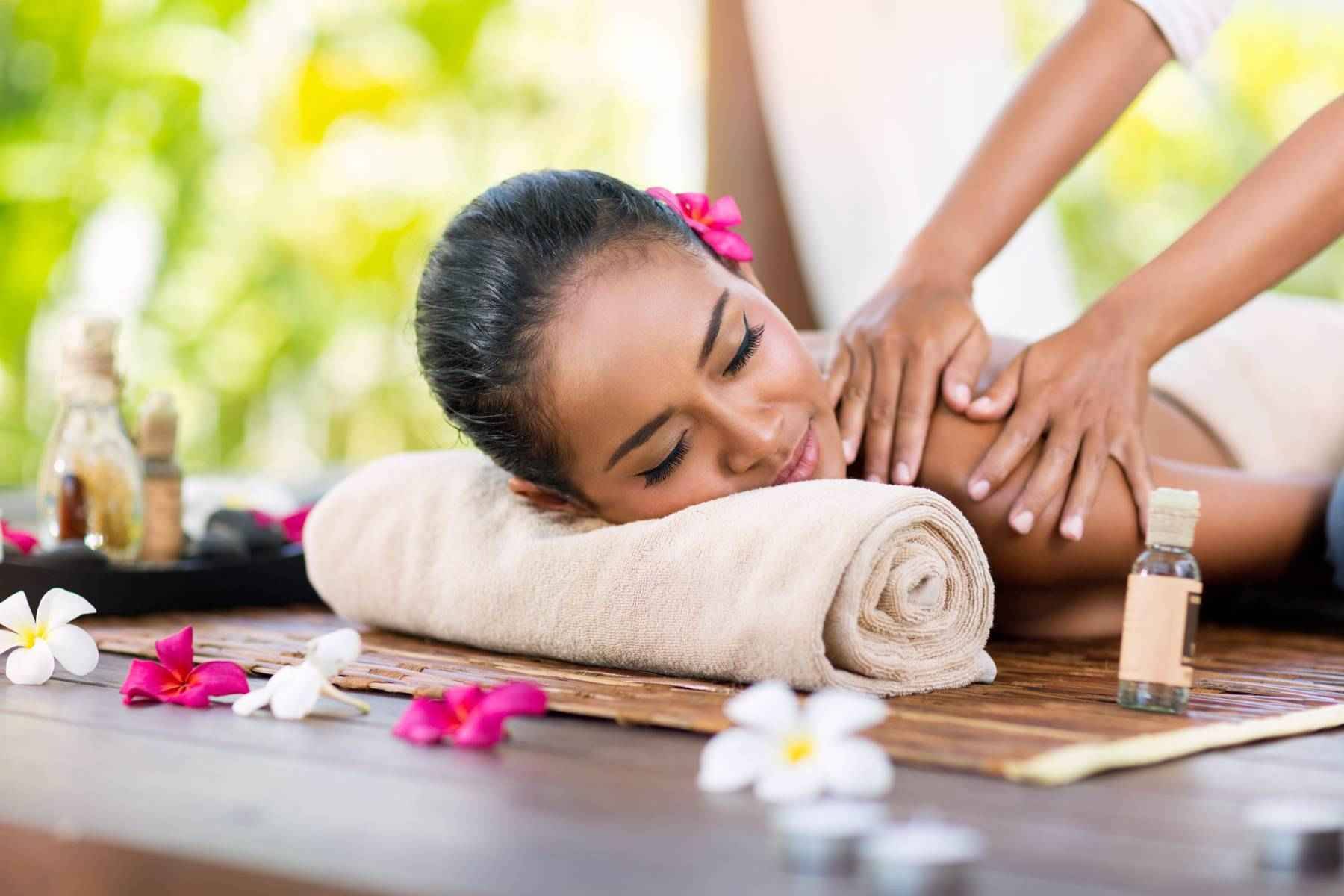 Smiling woman receiving a shoulder massage on a rolled towel, surrounded by flower petals and essential oils in a peaceful spa setting.
