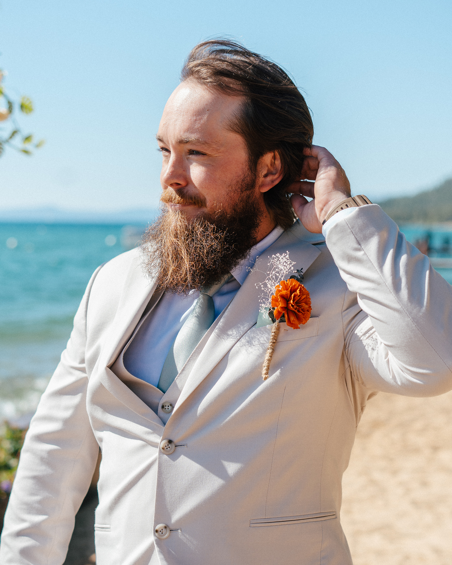 The groom adjusts his hair with Lake Tahoe glistening behind him during his beach wedding.
