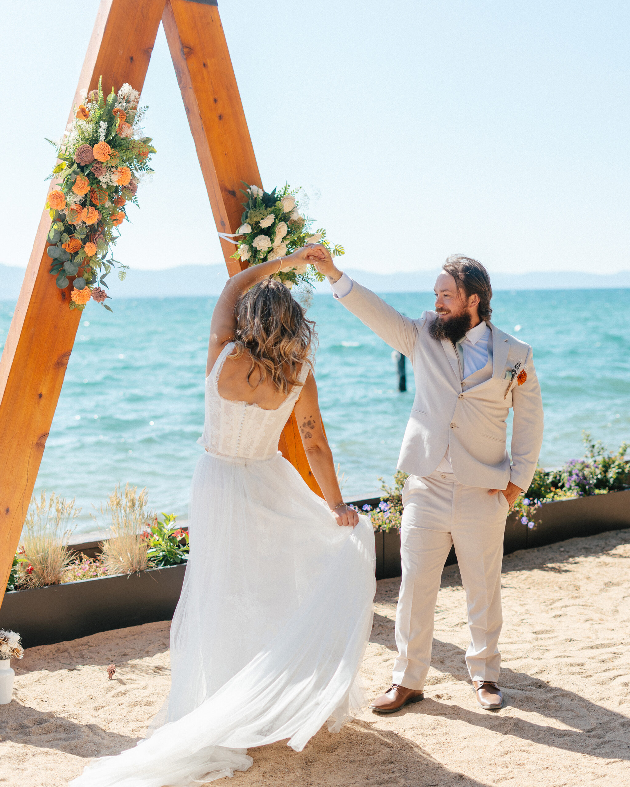 A newlywed couple shares their first dance beneath the floral arch by the shore.