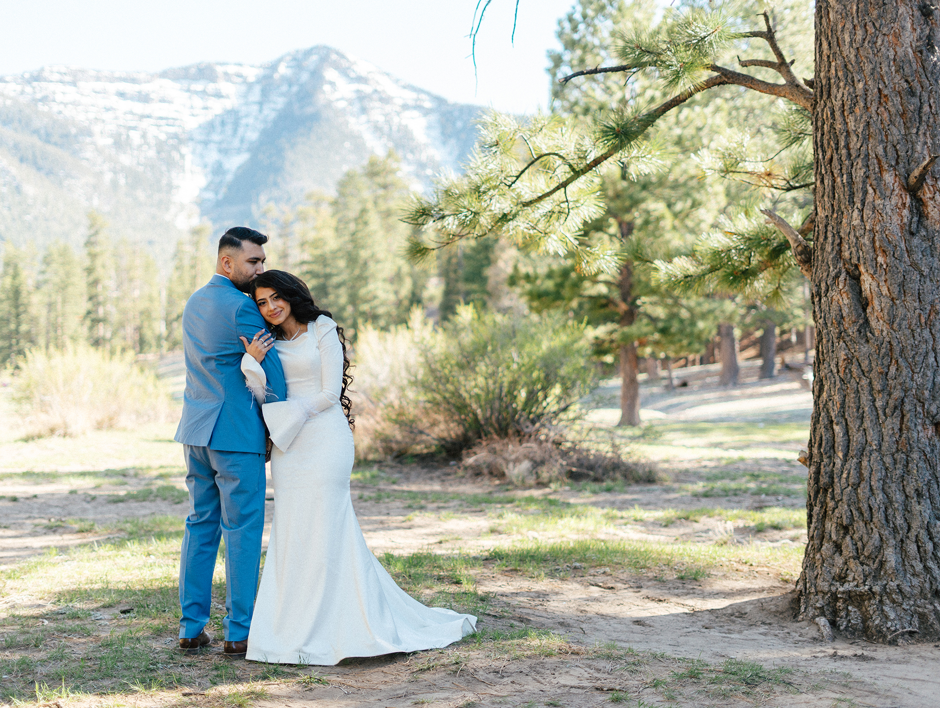 Bride and groom embrace under pine trees with snowy mountain backdrop at Lee Canyon.